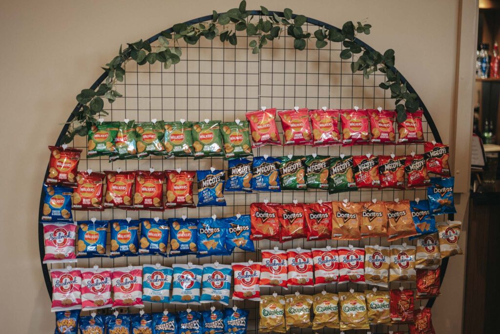 A circular black metal grid display brimming with colorful snack bags, including chips from Walkers and Doritos, leans elegantly against a plain beige wall at Stallingborough Grange. Greenery drapes over the grid, adding a decorative touch perfect for celebrating the bride's special day. © Aimee Lince Photography - Wedding photographer in Lincolnshire, Yorkshire & Nottinghamshire