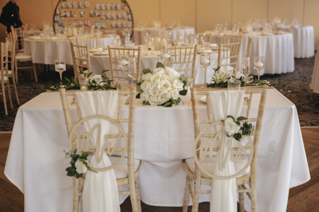 An elegant wedding reception at Stallingborough Grange features white linen-covered tables adorned with cream rose centerpieces and green foliage. Decorative wooden "Mr." and "Mrs." signs hang on chairs draped in white fabric. Soft lighting highlights this intimate setting for the bride's special day. © Aimee Lince Photography - Wedding photographer in Lincolnshire, Yorkshire & Nottinghamshire