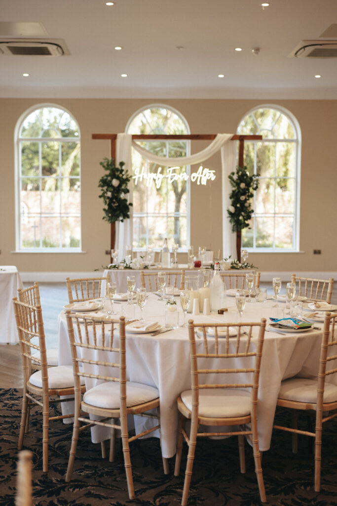A decorated wedding reception venue at Stallingborough Grange features round tables adorned with white tablecloths. Champagne glasses and cutlery are elegantly set, and a backdrop with greenery and a "Happily Ever After" sign graces the room. Large arched windows offer tree-filled views, bathing the space in natural light. © Aimee Lince Photography - Wedding photographer in Lincolnshire, Yorkshire & Nottinghamshire