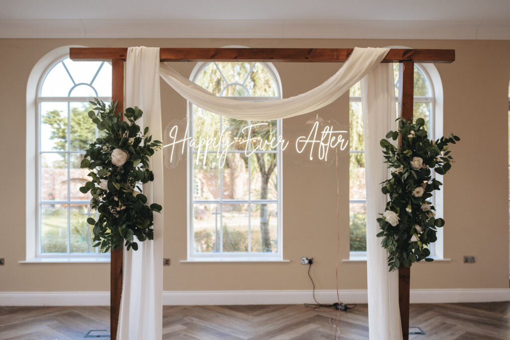 A wooden wedding arch at Stallingborough Grange, adorned with green foliage and white flowers, welcomes guests. Sheer white fabric drapes gracefully over the top, while a neon "Happy Ever After" sign shines in the center. Behind it, large windows reveal lush outdoor greenery and a wooden herringbone floor. © Aimee Lince Photography - Wedding photographer in Lincolnshire, Yorkshire & Nottinghamshire