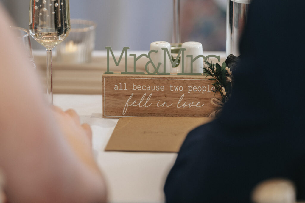A wedding table decoration at Stallingborough Grange features a wooden sign that reads "Mr & Mrs all because two people fell in love" in front of a mirror. A glass of champagne and salt and pepper shakers are also visible, with the romantic sign being the focal point amidst the blurred foreground. © Aimee Lince Photography - Wedding photographer in Lincolnshire, Yorkshire & Nottinghamshire