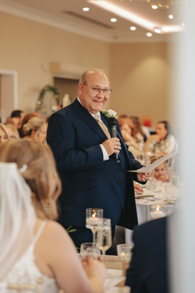 A bald man in a dark suit with a white rose boutonniere is speaking into a microphone, smiling warmly. He holds a paper while wedding guests at Stallingborough Grange sit at candlelit round tables. In the foreground, the bride with flowing hair and veil is partially visible, adding to the joy of the moment. © Aimee Lince Photography - Wedding photographer in Lincolnshire, Yorkshire & Nottinghamshire