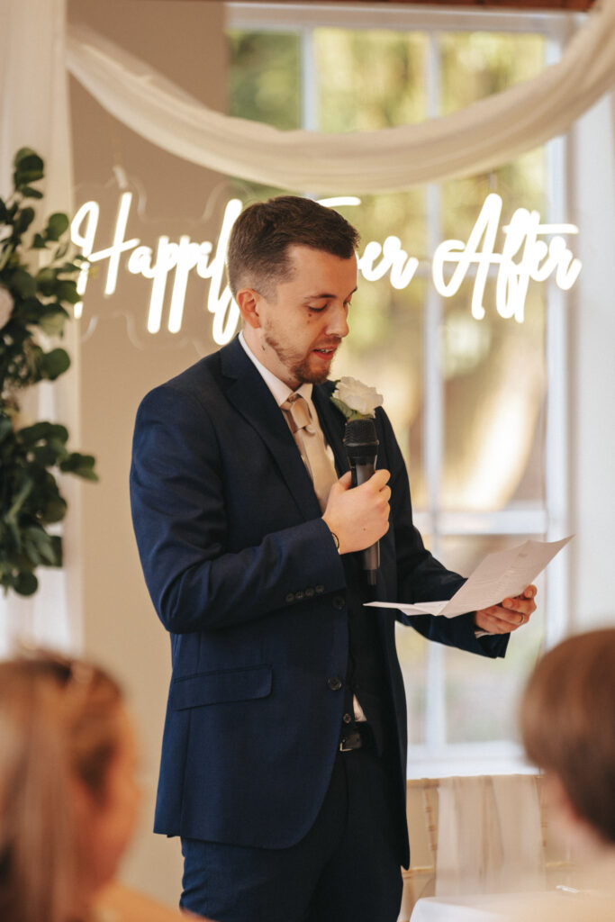 A man in a dark blue suit and beige tie holds a microphone and papers, speaking at an indoor wedding reception at Stallingborough Grange. A neon "Happily Ever After" sign glows in the background above white drapery, with a blurred audience eagerly waiting for the bride in the foreground. © Aimee Lince Photography - Wedding photographer in Lincolnshire, Yorkshire & Nottinghamshire
