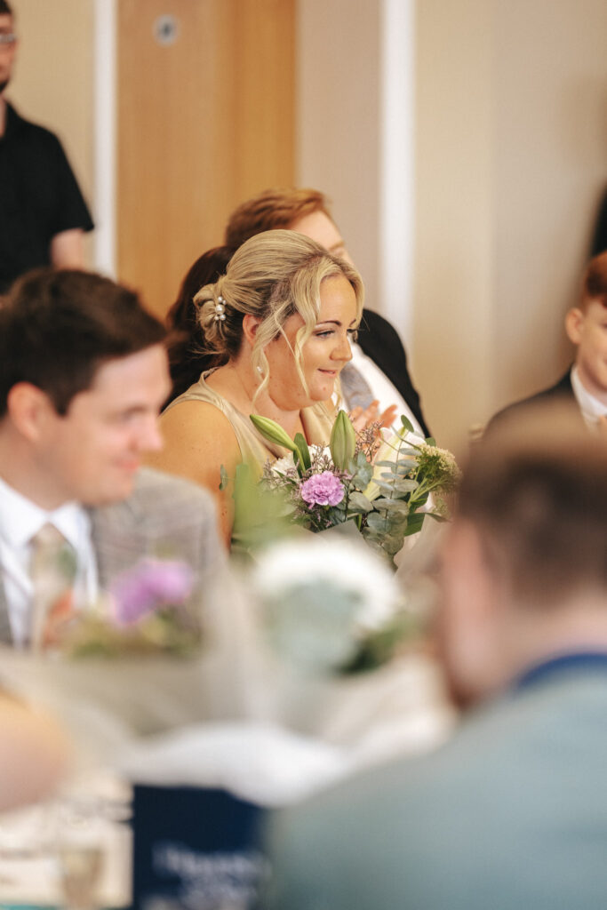 A woman with blonde hair, possibly the bride, is sitting at a table at Stallingborough Grange, smiling and holding a bouquet of purple and white flowers. She is wearing a light green dress, surrounded by several blurred guests. The setting appears to be indoors at a formal event or celebration. © Aimee Lince Photography - Wedding photographer in Lincolnshire, Yorkshire & Nottinghamshire