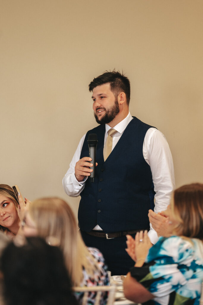 A man in a blue suit vest and beige tie speaks into a microphone at what seems to be a wedding. Seated guests listen attentively; a woman holding a smartphone possibly records the moment. Another guest claps amid the formal setting of Stallingborough Grange, framed by a neutral beige background. © Aimee Lince Photography - Wedding photographer in Lincolnshire, Yorkshire & Nottinghamshire