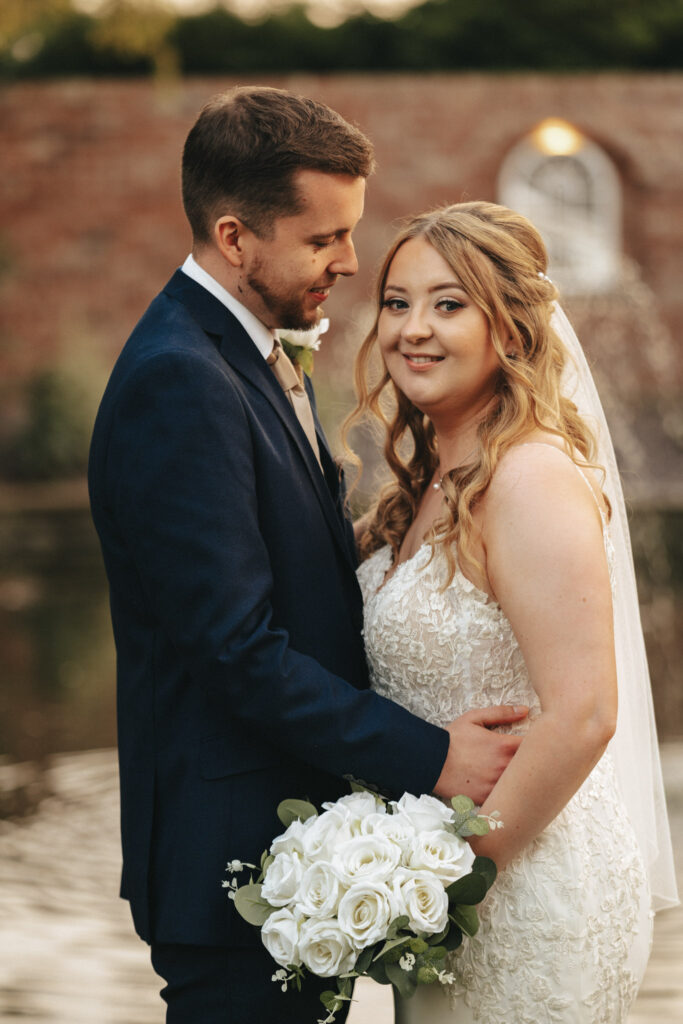 A bride and groom stand by a pond at Stallingborough Grange, with a brick wall and lush greenery in the background. The groom, in a dark suit, hugs the bride from the side. She wears a lacy white dress and holds a bouquet of white roses, smiling serenely at the camera. © Aimee Lince Photography - Wedding photographer in Lincolnshire, Yorkshire & Nottinghamshire