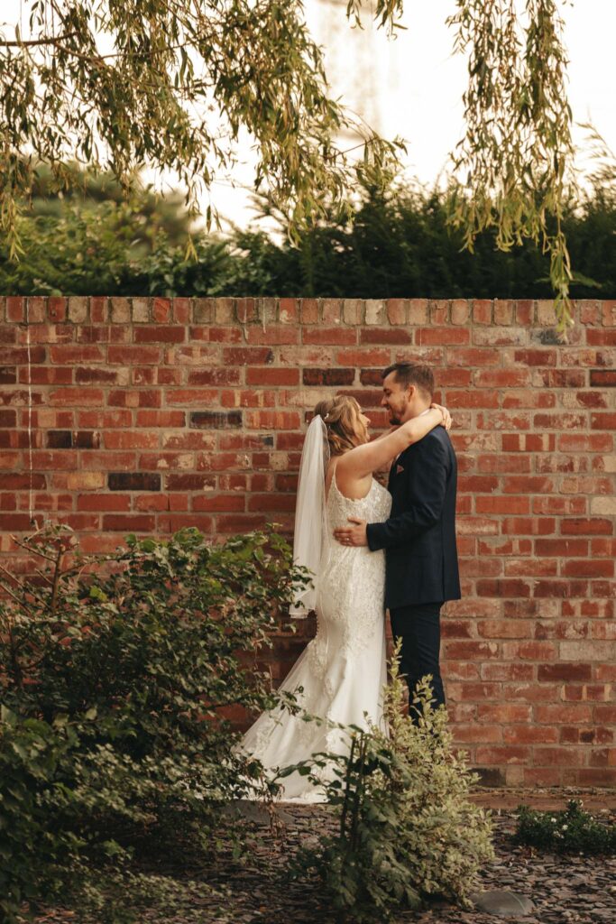The bride, in a white gown and veil, embraces the groom in his dark suit under a leafy tree at Stallingborough Grange. They smile at each other against a rustic red brick wall, surrounded by lush green shrubs and plants, creating a romantic wedding setting. © Aimee Lince Photography - Wedding photographer in Lincolnshire, Yorkshire & Nottinghamshire