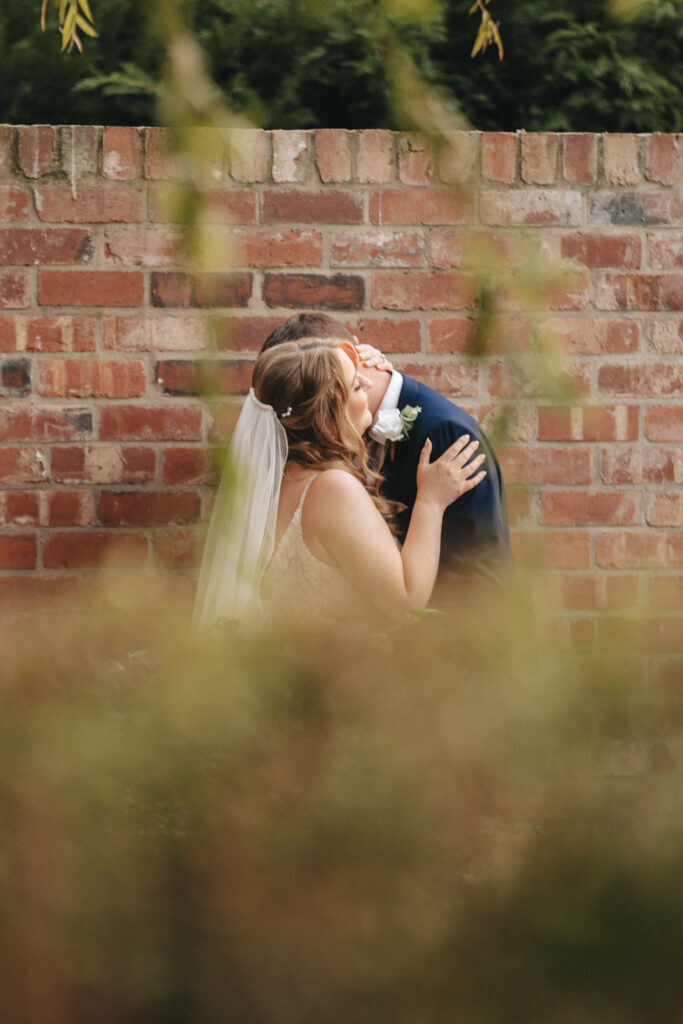 A bride and groom share a kiss in front of a rustic brick wall, capturing the magic of their wedding day. The bride's white dress and veil contrast beautifully with the groom's dark suit. Greenery partially frames the scene, adding a romantic touch to this Stallingborough Grange moment. © Aimee Lince Photography - Wedding photographer in Lincolnshire, Yorkshire & Nottinghamshire