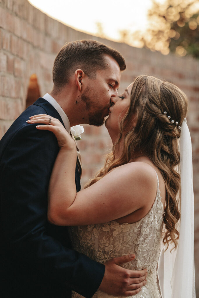 A bride and groom share a romantic kiss against a brick wall in the warm, soft glow of sunset. She wears a lace wedding dress with pearl hairpins, while he sports a white rose boutonniere. The intimate scene at Stallingborough Grange captures their love during this special moment. © Aimee Lince Photography - Wedding photographer in Lincolnshire, Yorkshire & Nottinghamshire