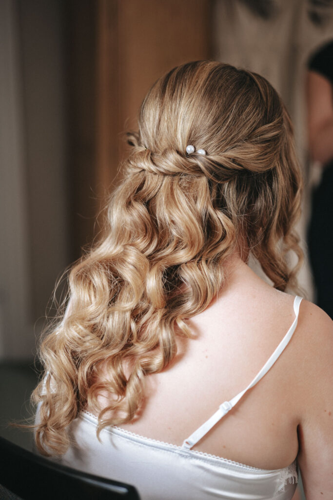 A bride with long, wavy blonde hair styled in loose curls, adorned with small white hairpins, sits with her back to the camera. She wears a white spaghetti strap top in an elegant interior setting, capturing a beautiful wedding hairstyle. © Aimee Lince Photography