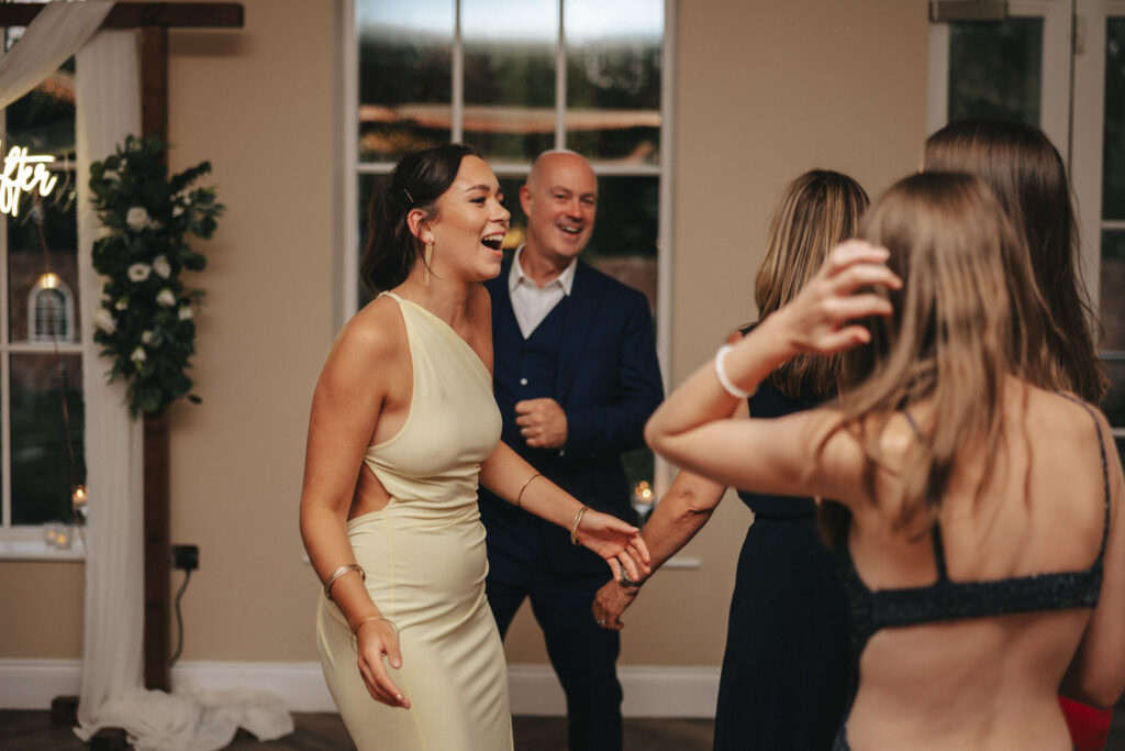 A lively group is joyfully dancing indoors. The bride, radiant in a light yellow dress, smiles as she holds hands with a bald man in a dark suit. In the foreground, another woman and a girl with long hair share the moment, while floral decorations hint at the romantic ambiance of Stallingborough Grange. © Aimee Lince Photography - Wedding photographer in Lincolnshire, Yorkshire & Nottinghamshire