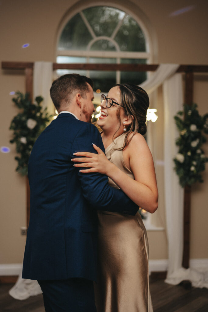 A couple is dancing closely, smiling at each other in a warmly lit room. The person on the left wears a navy blue suit, while the bride in a silky beige dress and glasses catches the light. A large window with white drapes and floral decorations hints at their wedding at Stallingborough Grange. © Aimee Lince Photography - Wedding photographer in Lincolnshire, Yorkshire & Nottinghamshire