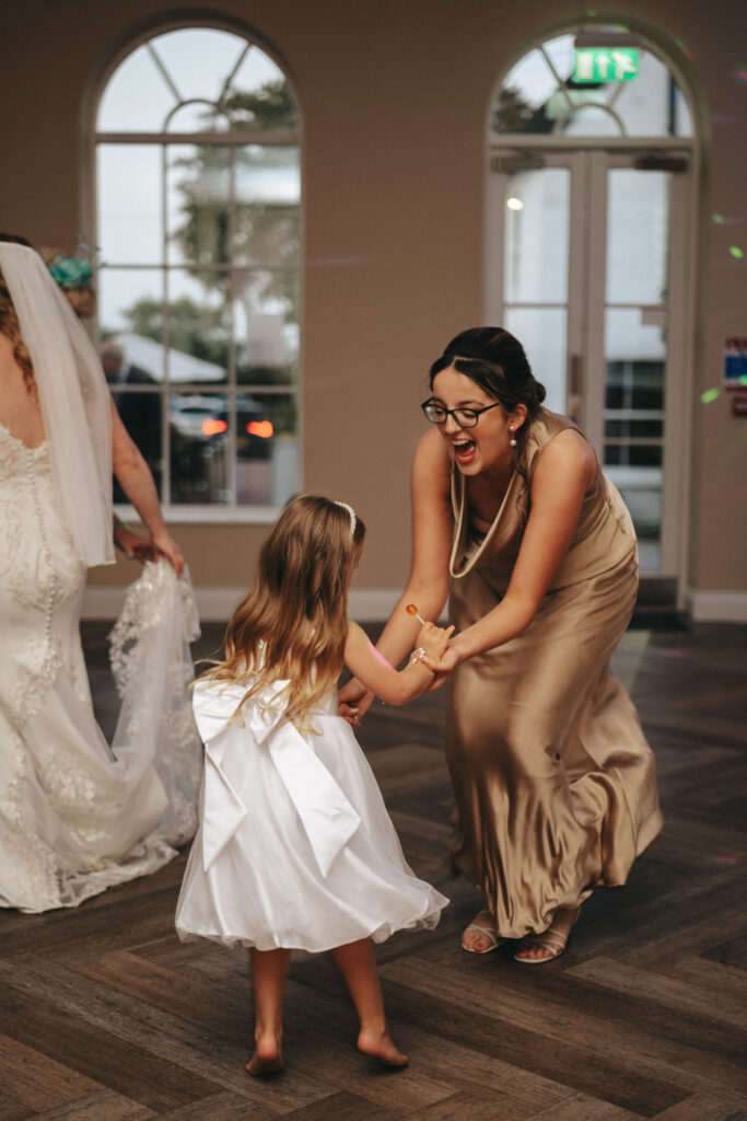 A woman in a gold dress and glasses joyfully dances with a young girl in a white dress with a large bow inside the sunlit room at Stallingborough Grange. The bride, radiant in her wedding dress and veil, watches them with a smile from the background. © Aimee Lince Photography - Wedding photographer in Lincolnshire, Yorkshire & Nottinghamshire