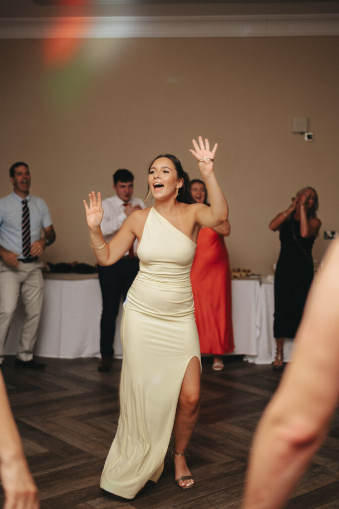 A person in a light-colored, one-shoulder dress dances energetically on a wooden floor at what seems to be a wedding celebration. She has one hand raised, wearing bracelets, and appears joyful. Other guests are visible in the background, dressed formally, clapping along at Stallingborough Grange. © Aimee Lince Photography - Wedding photographer in Lincolnshire, Yorkshire & Nottinghamshire
