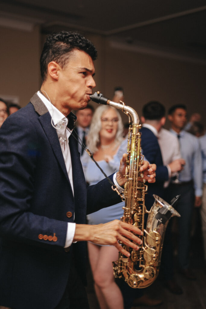 At a lively indoor event at Stallingborough Grange, a man in a navy blazer passionately plays the saxophone. A crowd, some holding drinks, gathers around him. The musician is focused and engaged, while the attendees soak in the enchanting performance, reminiscent of a joyous wedding celebration. © Aimee Lince Photography - Wedding photographer in Lincolnshire, Yorkshire & Nottinghamshire