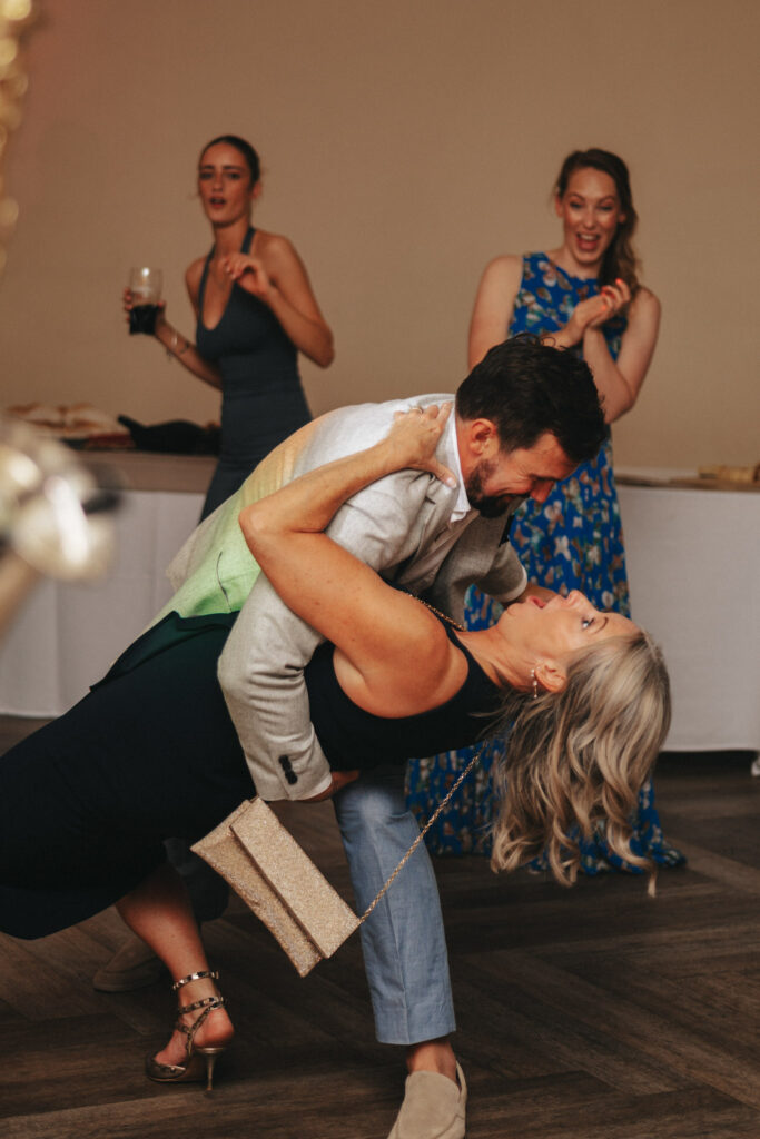 At a joyous wedding at Stallingborough Grange, a man in a light suit passionately dips the bride in her elegant black dress on the dance floor. She holds a sparkling clutch, smiling joyfully as three guests in the background display delighted expressions, capturing the lively atmosphere. © Aimee Lince Photography - Wedding photographer in Lincolnshire, Yorkshire & Nottinghamshire