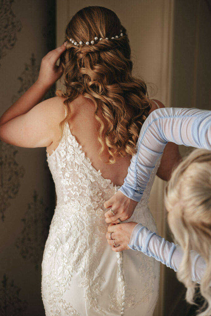 In the softly lit room with elegant wallpaper, a bride with wavy auburn hair, adorned with a pearl headpiece, stands as another person in a blue, long-sleeved dress fastens the buttons on her intricate lace wedding gown at Stallingborough Grange. © Aimee Lince Photography