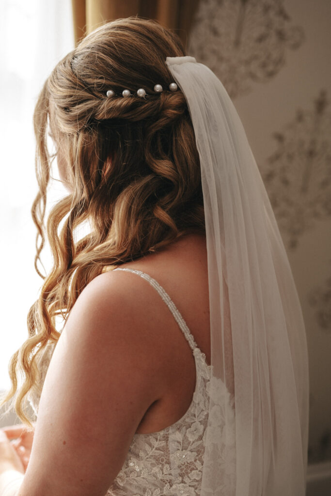 A bride in a lace wedding dress faces away, her long, wavy brown hair styled with a pearl hairpiece and veil. Soft light from a window illuminates her, revealing floral-patterned wallpaper in the background at Stallingborough Grange, creating an elegant and serene atmosphere. © Aimee Lince Photography