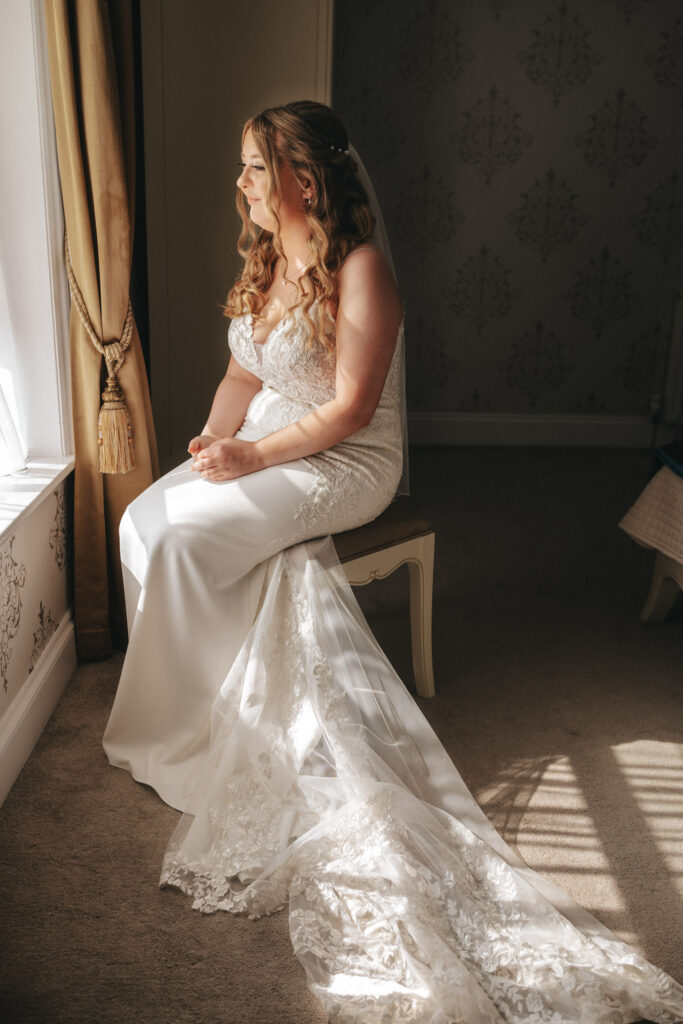 A bride in a white lace wedding dress with a long train sits on a stool, gazing out of the sunlit window at Stallingborough Grange. Her long, wavy hair cascades over her shoulders. The room, with its patterned wallpaper and gold curtains, creates a soft and serene atmosphere. © Aimee Lince Photography