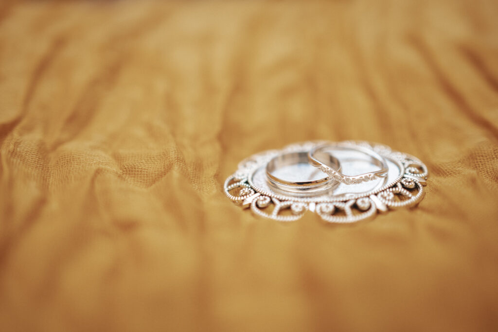 Two silver rings rest on an ornate, circular silver tray with detailed patterns. Set against the soft, golden fabric is a warm and elegant contrast to the metallic sheen of the rings and tray—a perfect backdrop for Emily & Luke's September wedding at Louth Town Hall. © Aimee Lince Photography