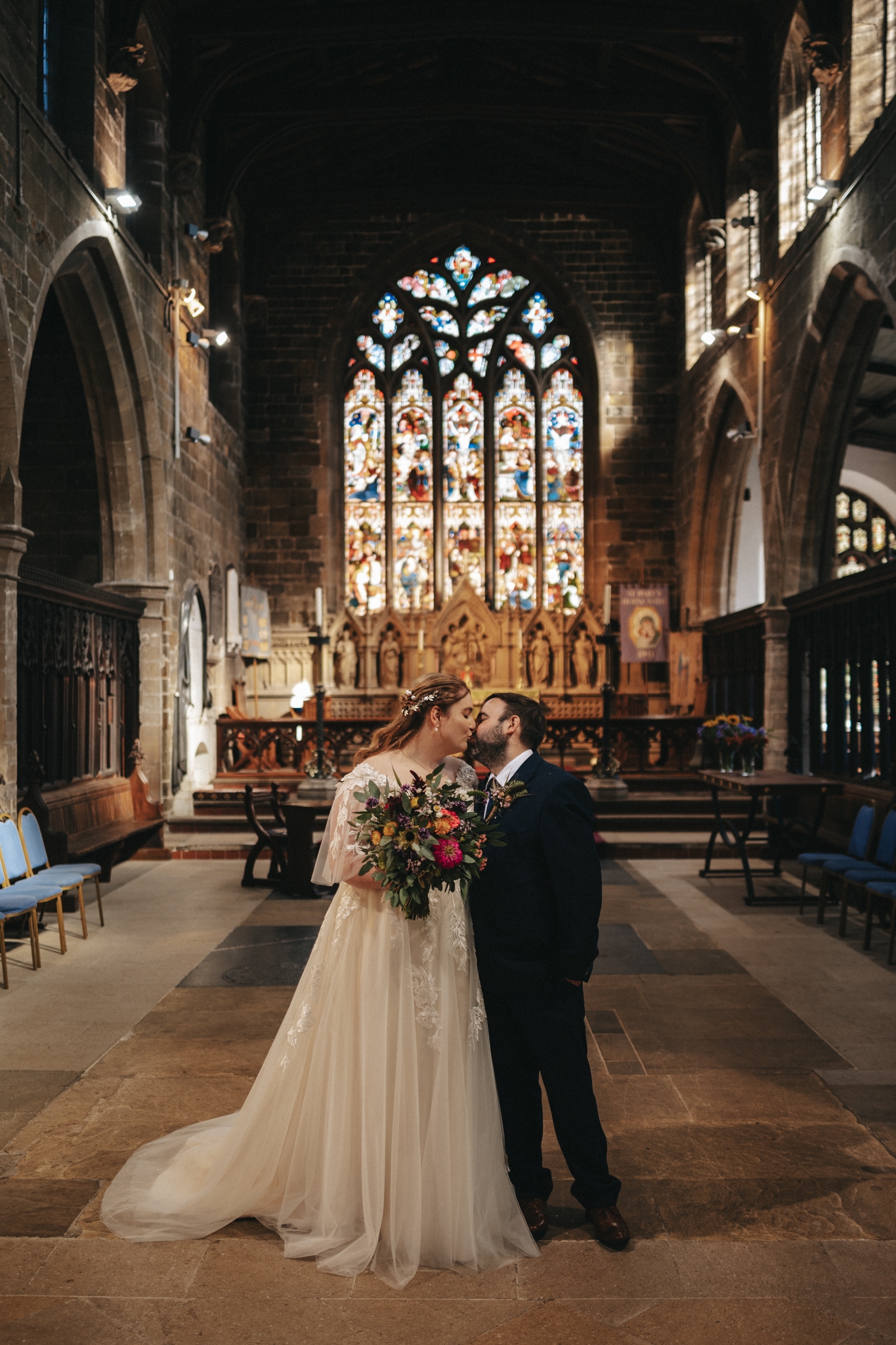 In a relaxed September wedding, Emily and Luke share a kiss in a grand church interior. The bride's elegant gown and the groom's dark suit complement their vibrant bouquet. Behind them, stained-glass windows and an ornate altar add to the romance of this special day. © Aimee Lince Photography