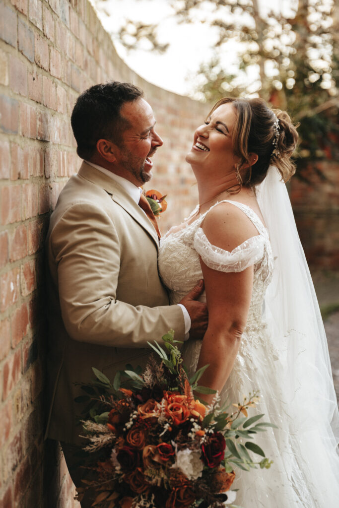 Kayleigh and Brett share a joyful moment against a brick wall at Stallingborough Grange Hotel. The bride, in a lace wedding dress and veil, holds an autumn wedding bouquet. The groom, in a light beige suit, stands close, smiling as they lovingly gaze at each other. The background is softly blurred. © Aimee Lince Photography