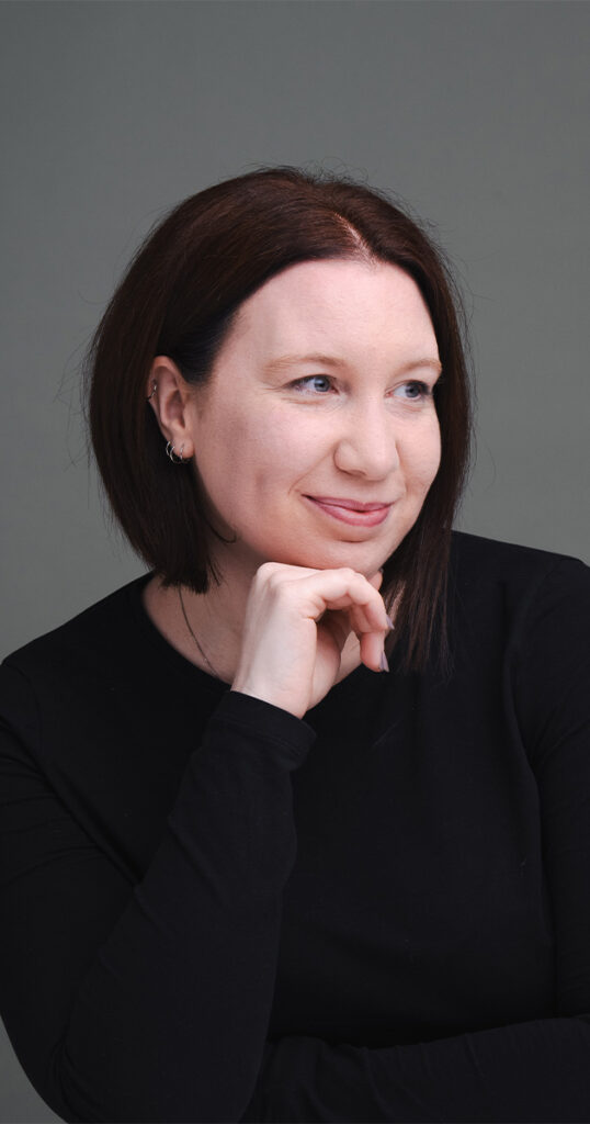 A person with shoulder-length dark hair is looking to the side with a slight smile, embodying a lifestyle vibe. They are wearing a black long-sleeve top, resting their chin on one hand. The backdrop is a plain, muted gray, adding an understated signature touch. © Aimee Lince Photography