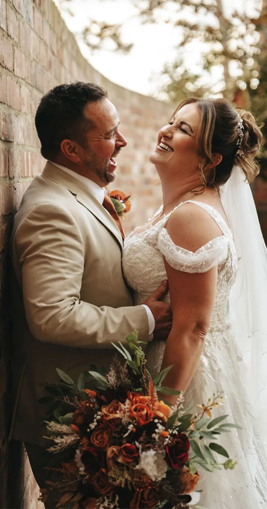 A bride and groom stand close, laughing together by a brick wall, embodying a charming autumn theme. The groom is in a beige suit, holding the bride's waist, while she wears a white lace gown and holds a bouquet of orange and white flowers. Blurred trees add a natural element to this delightful scene. © Aimee Lince Photography - Wedding photographer in Lincolnshire, Yorkshire & Nottinghamshire