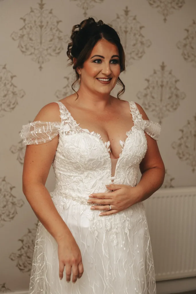 A smiling woman in a white, off-shoulder lace wedding dress stands indoors against patterned wallpaper at Stallingborough Grange. Her dark hair is styled up with subtle makeup. Her right hand rests on her waist, showcasing a sparkling ring that complements the elegant theme of the occasion. © Aimee Lince Photography - Wedding photographer in Lincolnshire, Yorkshire & Nottinghamshire