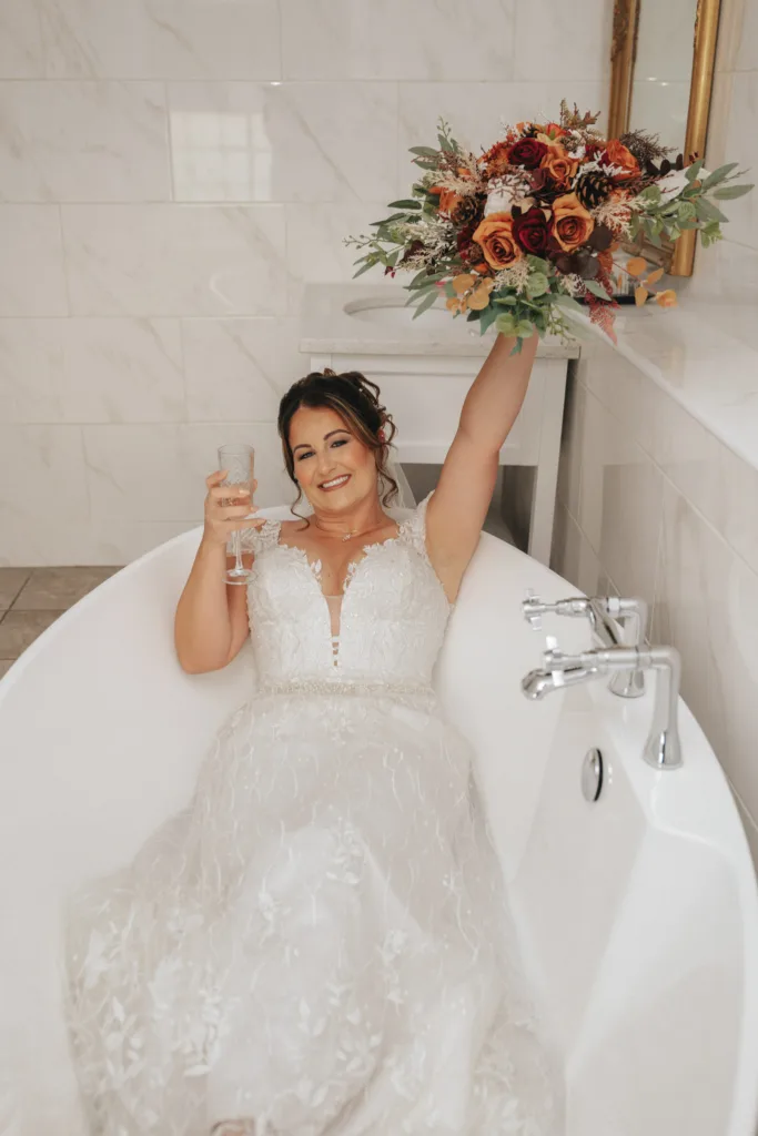 The bride in a white lace wedding dress smiles, lounging in a white bathtub. Embracing the autumn theme, she raises a bouquet of red, orange, and green flowers with one hand and holds a champagne glass in the other. The bathroom features white tiled walls and a faucet beside her in the tub. © Aimee Lince Photography - Wedding photographer in Lincolnshire, Yorkshire & Nottinghamshire