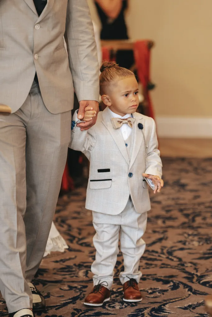 A young child in a light gray suit with a bow tie holds the hand of an adult in a matching ensemble at an elegant autumn wedding. Their brown shoes echo the season's theme as they walk on a blue-patterned carpet, lending an air of sophistication to the Stallingborough Grange ceremony. © Aimee Lince Photography - Wedding photographer in Lincolnshire, Yorkshire & Nottinghamshire