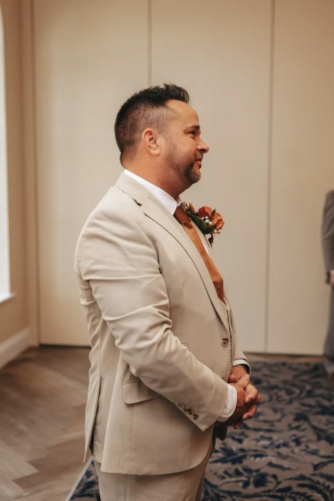 A man in a light beige suit with a white shirt and brown tie stands smiling, hands clasped in front, evoking an autumn theme. His short dark hair and beard complement the boutonniere with brown flowers on his lapel. He poses gracefully against a neutral wall and patterned blue carpet at Stallingborough Grange. © Aimee Lince Photography - Wedding photographer in Lincolnshire, Yorkshire & Nottinghamshire