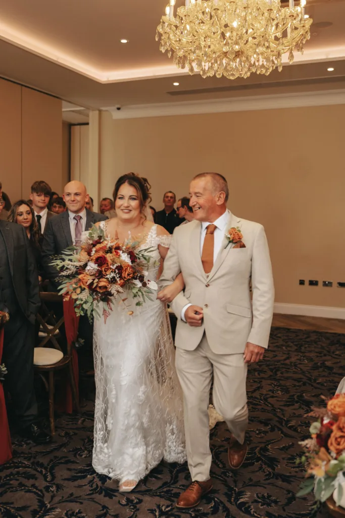 A bride in a lace gown walks down the aisle with an older man in a beige suit, holding a vibrant bouquet that complements the autumn theme. Guests watch them in the warmly lit room of Stallingborough Grange, where a crystal chandelier adds to the joyful and celebratory atmosphere. © Aimee Lince Photography - Wedding photographer in Lincolnshire, Yorkshire & Nottinghamshire
