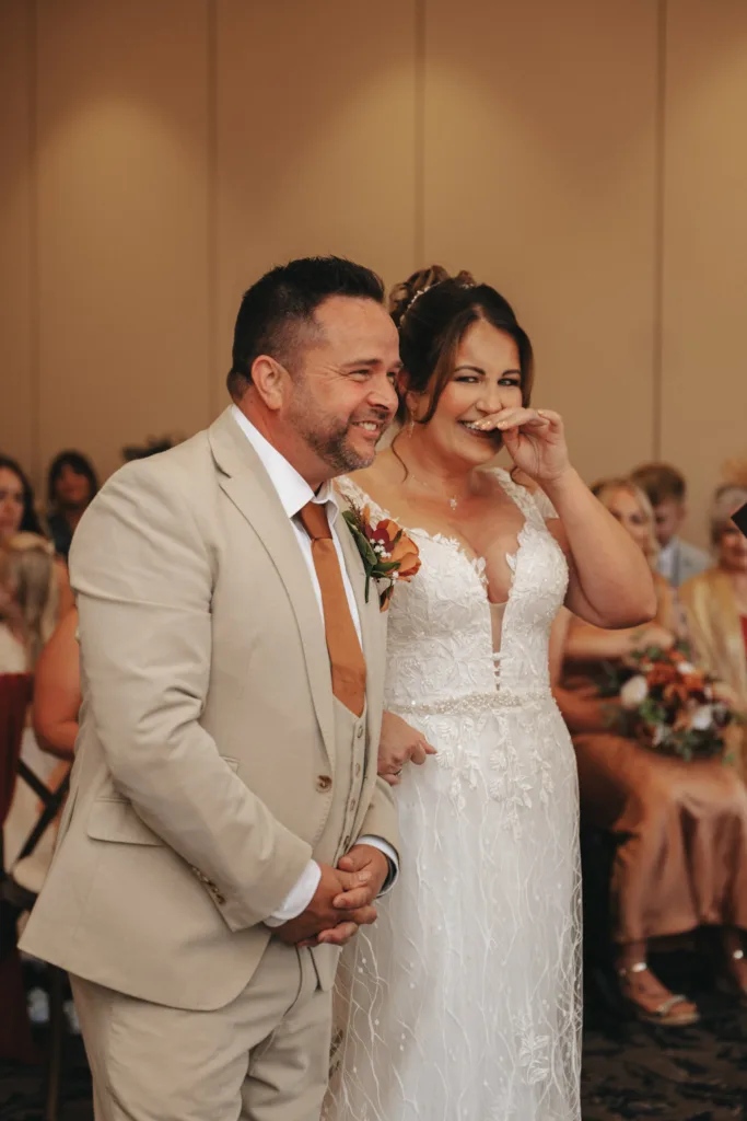A couple stands at their wedding ceremony at Stallingborough Grange Hotel. The groom wears a beige suit with a burnt orange tie, smiling warmly. The bride, in a white dress with lace details, holds a hand to her mouth, appearing emotional as guests in soft tones sit behind them in the gently lit room. © Aimee Lince Photography - Wedding photographer in Lincolnshire, Yorkshire & Nottinghamshire