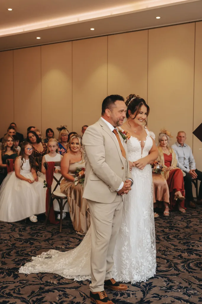 A bride in a lace gown and a groom in a beige suit stand smiling at their autumn-themed wedding ceremony at Stallingborough Grange. Seated guests watch from behind, including a child in a white dress. The room has beige walls and patterned carpet, with bouquets and warm lighting enhancing the festive atmosphere. © Aimee Lince Photography - Wedding photographer in Lincolnshire, Yorkshire & Nottinghamshire