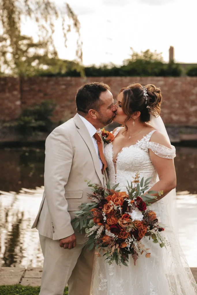 In an enchanting autumn wedding scene, a couple shares a kiss in front of a pond, framed by a brick wall and lush greenery. The bride dazzles in her white lace gown with a bouquet of rust, orange, and white flowers. The groom complements her in a beige suit with a rust-colored tie. © Aimee Lince Photography - Wedding photographer in Lincolnshire, Yorkshire & Nottinghamshire