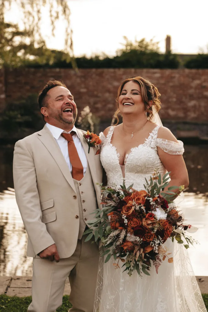 A joyful couple stands by a serene pond at Stallingborough Grange. The man wears a light beige suit with a rust-colored tie, laughing heartily. The woman, in a detailed white lace dress, holds an autumn bouquet of rust, cream, and green flowers, smiling warmly against the brick wall and lush foliage backdrop. © Aimee Lince Photography - Wedding photographer in Lincolnshire, Yorkshire & Nottinghamshire