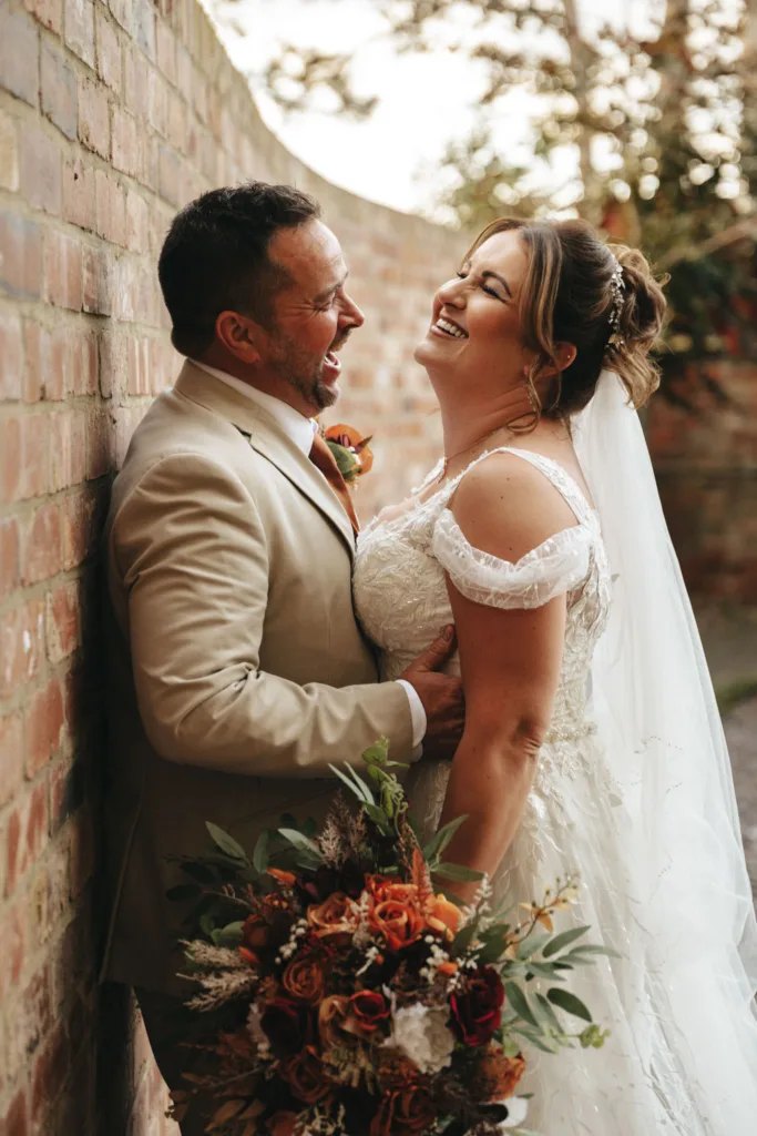 A bride and groom are joyfully laughing together against a brick wall in the soft glow of autumn light. The bride wears a white lace wedding dress, holding a bouquet of orange and white flowers, complementing their fall theme. The groom is in a light beige suit as they gaze lovingly at each other. © Aimee Lince Photography - Wedding photographer in Lincolnshire, Yorkshire & Nottinghamshire