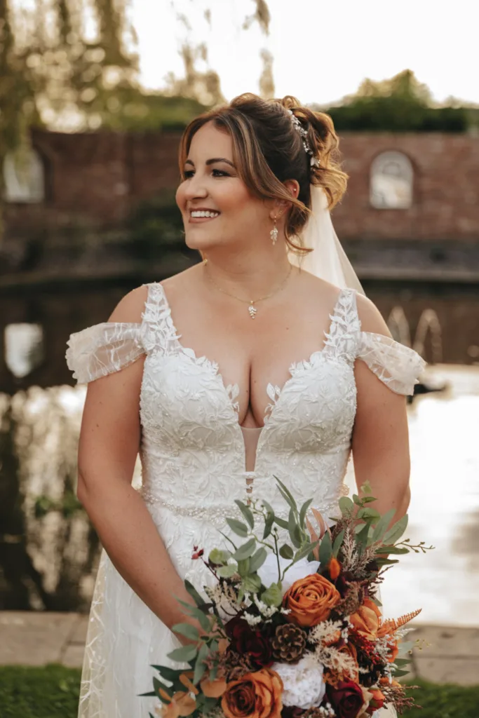 A bride stands outdoors near a pond, smiling and holding a bouquet of orange and white flowers, perfectly capturing the autumn wedding theme. Her white lace dress has off-the-shoulder sleeves and floral embroidery. Her hair is in an updo with loose curls, complemented by a veil and drop earrings. A brick wall is in the background. © Aimee Lince Photography - Wedding photographer in Lincolnshire, Yorkshire & Nottinghamshire