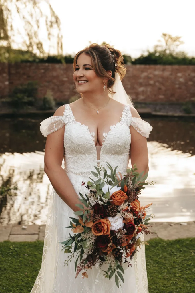 A bride stands in front of a pond at Stallingborough Grange Hotel, smiling while holding a bouquet with red and white flowers, greenery, and brown accents. Her white dress features lace detailing and off-the-shoulder sleeves. Her hair is styled up with a veil. The backdrop hints at an enchanting autumn wedding with its brick wall and trees. © Aimee Lince Photography - Wedding photographer in Lincolnshire, Yorkshire & Nottinghamshire