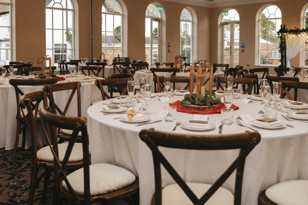 A banquet hall at Stallingborough Grange is set for an autumn-themed event, featuring round tables with white tablecloths and wooden chairs. Each table boasts white napkins, glasses, cutlery, and centerpieces of lanterns and greenery on red runners. Arched windows bring in natural light over the carpeted floor. © Aimee Lince Photography - Wedding photographer in Lincolnshire, Yorkshire & Nottinghamshire