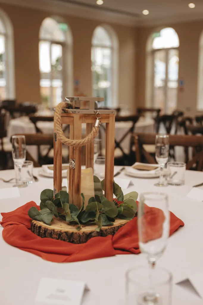 A rustic autumn wedding centerpiece features a wooden lantern with a lit candle inside, surrounded by greenery and set on a wooden slice. Resting on burnt orange cloth, this charming display at Stallingborough Grange includes champagne glasses and folded cards, with chairs elegantly arranged in the background. © Aimee Lince Photography - Wedding photographer in Lincolnshire, Yorkshire & Nottinghamshire