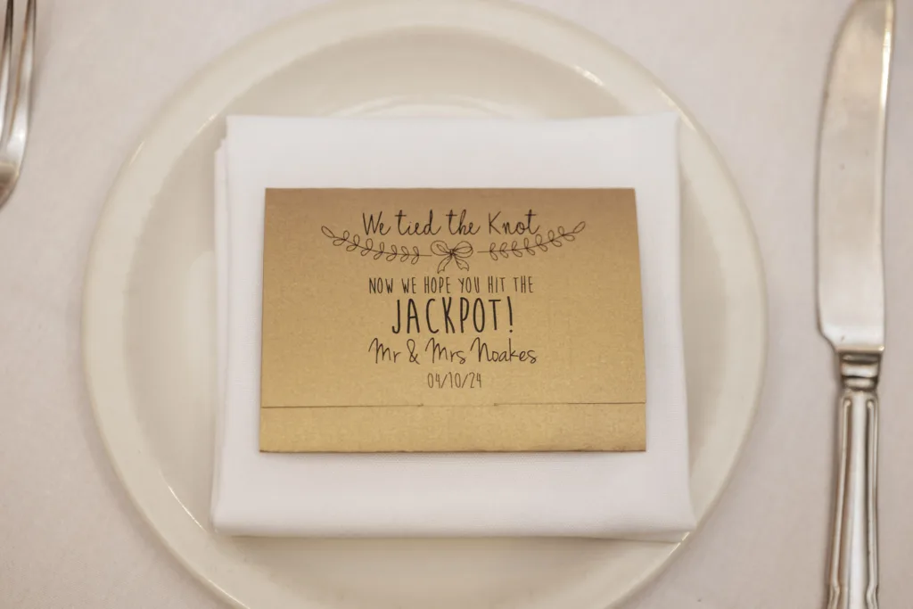 A wedding place setting at the elegant Stallingborough Grange Hotel features a white plate and silver cutlery. A gold card atop an autumn-themed napkin reads, “We tied the knot! Now we hope you hit the jackpot! Mr. & Mrs. Noakes 09/10/14.” The napkin is neatly folded with cutlery on either side. © Aimee Lince Photography - Wedding photographer in Lincolnshire, Yorkshire & Nottinghamshire