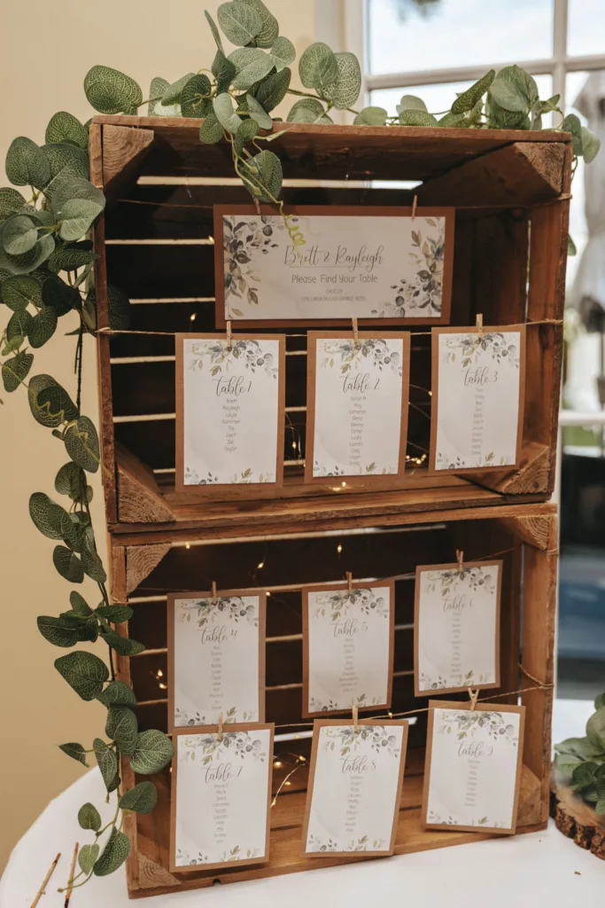 Two wooden crates stacked, with greenery draped around, set the stage for an elegant autumn wedding. Ten cards attached with clothespins list seating arrangements for tables 1-10. A sign above reads, "Please Find Your Table." The setup at Stallingborough Grange Hotel features a window in the background. © Aimee Lince Photography - Wedding photographer in Lincolnshire, Yorkshire & Nottinghamshire