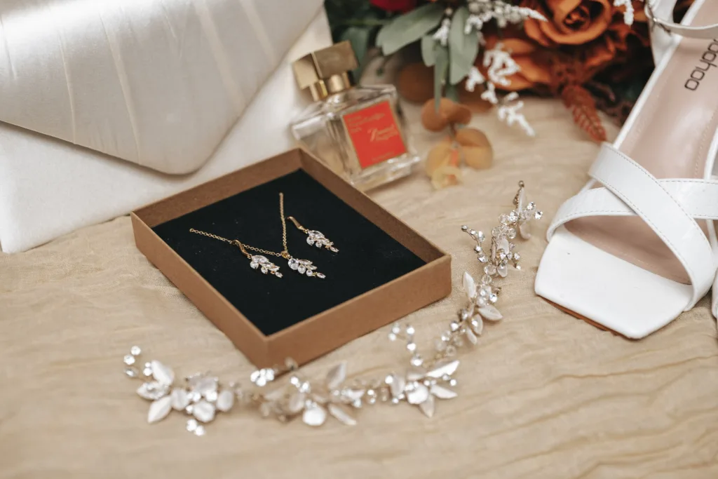 A flat lay of bridal accessories: delicate gold and silver earrings in a box, a bottle of perfume, a white high-heeled shoe, and a floral hairpiece. The soft beige fabric complements hints of orange flowers, setting an autumnal mood at Stallingborough Grange Hotel. © Aimee Lince Photography - Wedding photographer in Lincolnshire, Yorkshire & Nottinghamshire