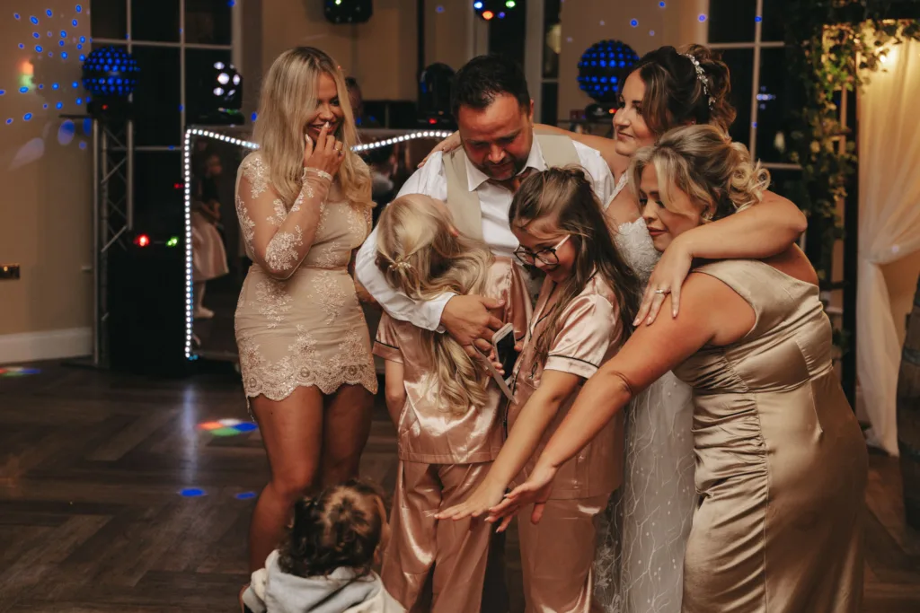 A joyful group at an autumn wedding, including a bride in a white dress and others in shiny attire, gather excitedly around a small child in pajamas. A man stands at the center, smiling. The setting features dim lighting, a lit stage backdrop with an autumn theme, and a wooden floor. © Aimee Lince Photography - Wedding photographer in Lincolnshire, Yorkshire & Nottinghamshire