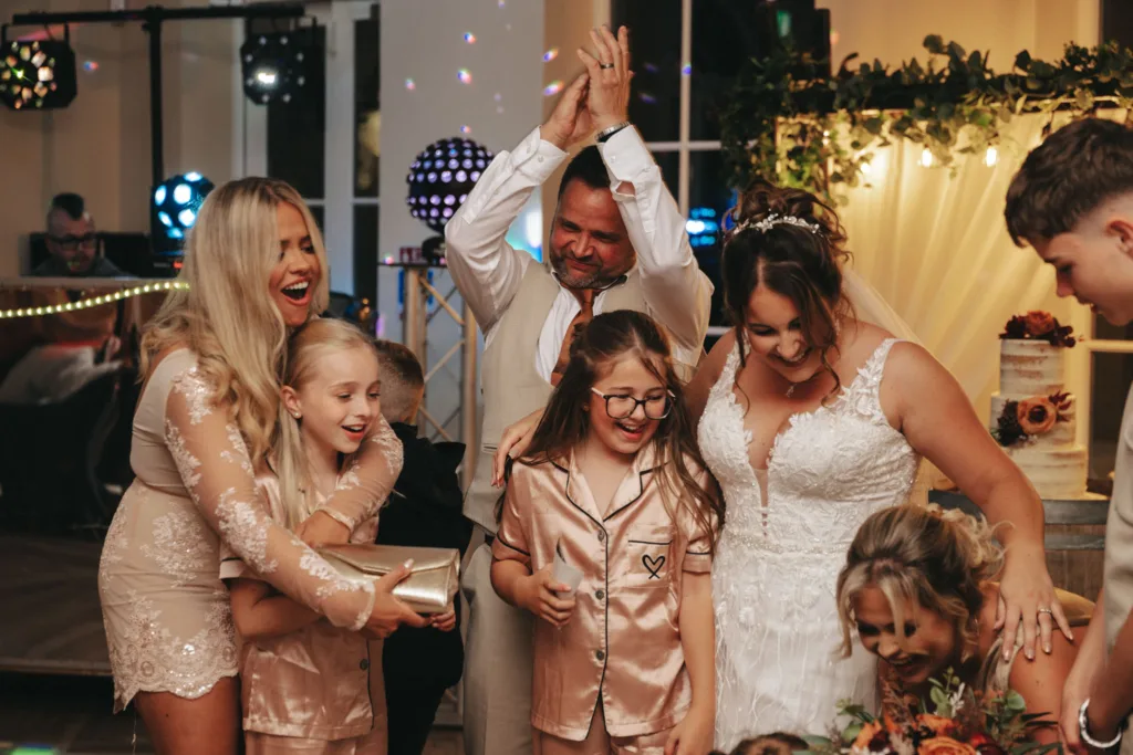 A joyful wedding celebration unfolds at the Stallingborough Grange Hotel, with a bride in a white gown surrounded by laughing guests. A man enthusiastically claps behind her. Two young girls in matching outfits, one with glasses, smile as they enjoy the festive atmosphere in this beautifully themed room adorned with lights and flowers. © Aimee Lince Photography - Wedding photographer in Lincolnshire, Yorkshire & Nottinghamshire