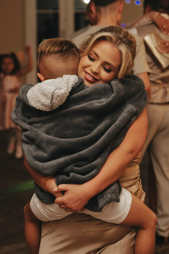At what seems to be an indoor event, possibly an autumn wedding at Stallingborough Grange, a blonde-haired woman smiles warmly as she embraces a child wrapped in a grey blanket. The child, with short hair and white shorts, is surrounded by guests, including one in a pink dress. © Aimee Lince Photography - Wedding photographer in Lincolnshire, Yorkshire & Nottinghamshire