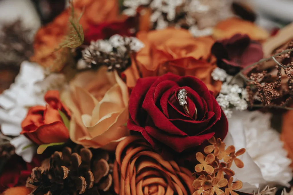 A bouquet with a variety of flowers in rich autumnal colors, including deep red, orange, and beige hues, echoing the rustic theme of Stallingborough Grange Hotel. A diamond engagement ring is nestled within a red rose amidst pinecones and brown dried foliage for a warm, inviting ambiance. © Aimee Lince Photography - Wedding photographer in Lincolnshire, Yorkshire & Nottinghamshire
