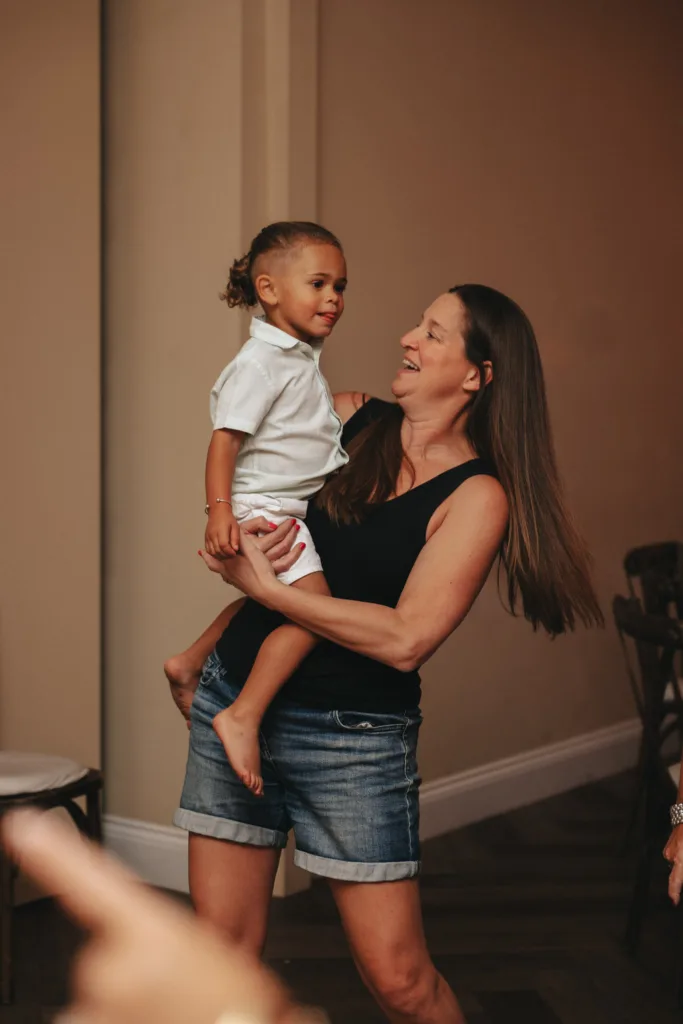 A woman with long hair, wearing a black tank top and denim shorts, holds a young child in a white shirt and shorts. Both are smiling joyfully. They are in a cozy room with beige walls and wooden flooring that echoes an autumn theme. A blurred arm is visible in the foreground on the left. © Aimee Lince Photography - Wedding photographer in Lincolnshire, Yorkshire & Nottinghamshire